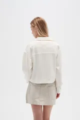 Chaqueta color blanco hueso de lino con cuello camisero, cierre frontal con botones marrones, bolsillos de parche en el pecho y corte holgado.