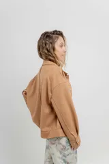 Chaqueta color beige de corte recto, estilo camisa, confeccionada en gamuza con elastano. Presenta cuello camisero, cierre frontal con botones metálicos, bolsillos grandes en el frente y charreteras en los hombros.