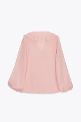 Camisa color rosa pálido confeccionada en tejido ligero con lyocell. Presenta escote en V, cierre frontal con botones, mangas largas abullonadas con puños elásticos y detalle de volantes en el frente.