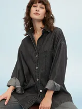 Camisa de denim negro con corte oversize, cuello clásico, cierre frontal con botones metálicos y bolsillo de parche en el pecho.
