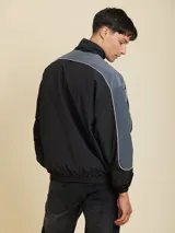 Campera rompevientos negra con mangas ranglan en gris denim y vivos blancos. Logo de la marca estampado en el pecho. Cierre frontal con cremallera y bolsillos laterales. Puños elastizados.