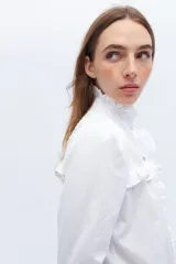 Camisa blanca de algodón con cuello alto con volados y bordado calado en el frente. Tiene mangas largas con puños abotonados y cierre frontal con botones.