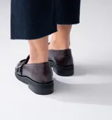 Mocasines de cuero color marrón con estampado de cocodrilo, detalle de hebilla metálica plateada y suela de goma negra. Diseño clásico con punta redonda y calce cómodo.