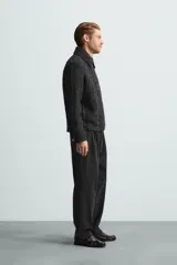 Sobrecamisa negra con textura de tejido bouclé y rayas verticales plateadas. Tiene cuello solapa, manga larga con puños abotonados, bolsillos laterales y cierre frontal con cremallera.