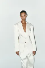 Blazer blanca de tejido satinado, con cuello solapa y escote pico, manga larga con hombreras, bolsillos con solapa en delantero y falso bolsillo en pecho, bajo con abertura en espalda y cierre frontal con botón.