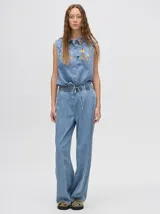 Mono largo de jean celeste con corte holgado, cintura elastizada con cordón ajustable, sin mangas, cuello camisero y cierre frontal con botones. Presenta bordados de motivos playeros en el pecho.