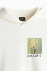 Buzo canguro color crema con capucha cruzada de doble capa, hombros caídos, bolsillo tipo canguro, puños y bajo acanalados. Estampado frontal con flores y texto "Beautanical". Corte holgado.