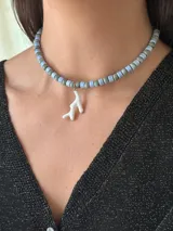 Collar corto tipo choker compuesto por cuentas cilíndricas de cerámica en tonos azul claro y gris jaspeado, con un dije colgante en forma de coral blanco esmaltado.