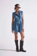 Vestido camisero corto de denim rígido liviano, color celeste, sin mangas, con acceso por botones en delantero y cinto con hebilla metálica plateada.
