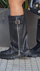 Botas de caña alta color negro, con suela track y cierre lateral con cremallera. Presentan una correa con hebilla alrededor de la caña.