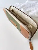 Billetera grande con cierre metálico, confeccionada en cuero con diseño de bloques de color: verde oliva texturizado, marrón claro liso y blanco/crema texturizado. Posee un monedero interno y un tirador de cuero marrón claro.