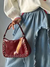Cartera tipo baguette color borgoña con tachas plateadas en los bordes y asa corta.
