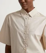 Camisa de tricolina color beige de la marca Cortelle, con cuello camisero y mangas cortas. Presenta un diseño con recortes frontales horizontales y cierre central con botones diferenciados.