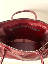 Bolso grande de cuero marrón con textura tipo cocodrilo, estructura rígida y asas para llevar al hombro. Tiene una tira con tachas y hebillas en cada extremo, también lleva lazos en los costados.