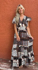 Vestido largo con estampado tipo patchwork en tonos tierra, blanco y azul oscuro. Presenta escote cruzado en V y mangas cortas. La falda es larga y con vuelo.