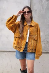 Campera estilo bomber o trucker de color mostaza/naranja quemado con efecto acid wash o desgastado. Presenta cuello camisero, cierre frontal y puños y cintura ajustados con elástico. Posee bolsillos frontales con solapa y botón.