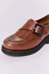 Zapato mocasín color marrón de cuero con hebilla metálica negra y detalle de flecos.