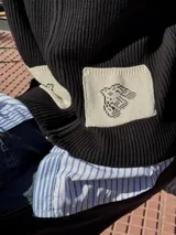 Campera tejida con textura vanizada a rayas horizontales en blanco y negro, con cierre frontal y flecos negros en forma de V en el pecho.