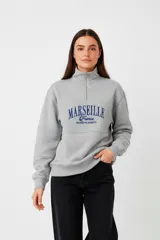 Buzo de algodón color beige claro (hielo) con cuello alto y cierre parcial frontal. Presenta un diseño holgado con franjas horizontales a la altura del pecho y texto estampado en color marrón oscuro.