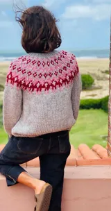 Sweater de punto grueso color gris claro con cuello redondo y diseño con motivos geométricos en tonos rosa y borgoña en la parte superior del pecho y hombros.