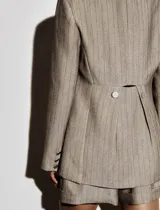 Blazer color beige con finas rayas verticales negras, solapas de muesca, cierre frontal con dos botones, bolsillos delanteros con solapa y botones, y botones decorativos en los puños.