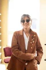 Blazer de cuero gamuza color marrón, con solapas y un botón frontal blanco. La prenda está decorada con varios pines metálicos y uno de ónix negro en la solapa izquierda.