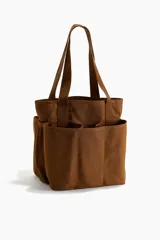 Bolso shopper marrón de lona de algodón con doble asa de mano y múltiples compartimentos exteriores, tres en los lados largos y uno de malla en los laterales.