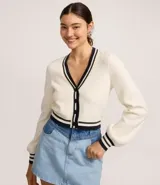 Cardigan corto de punto color crema con escote en V ribeteado en negro y blanco. Presenta cierre frontal de botones negros y puños con el mismo ribete a rayas. Las mangas son largas y ligeramente abullonadas.