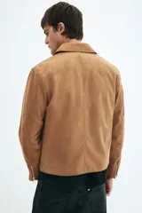 Campera de gamuza color camel, con cuello camisero, cierre frontal y bolsillos laterales con cierre. Presenta un corte estándar y forro interior.