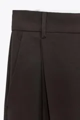Pantalón marrón de tiro alto con trabillas, doble pliegue, bolsillos laterales y de vivo en la espalda, pernera ancha y cierre frontal con cremallera y gancho metálico.