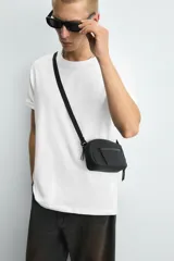 Bandolera negra de acabado engomado con correa ajustable y extraíble. Tiene un compartimento principal con cierre de cremallera, un bolsillo interior con cremallera y un bolsillo frontal con cremallera.