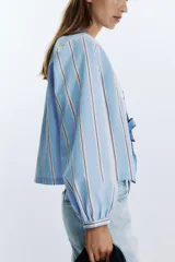 Camisa celeste con rayas verticales marrones y blancas, cuello redondo con escote en pico y mangas largas abullonadas. Cierre frontal con lazos con pespuntes marcados.