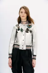 Campera bomber plateada de cuero ovino con cuello, puños y ruedo acanalados con lurex. Presenta cierre frontal con botones a presión y apliques de parches bordados con lentejuelas y mostacillas. Forro de poliéster.