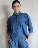 Camisa de jean azul con cuello con volados, mangas 3/4 abullonadas y cierre frontal con botones.