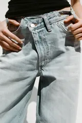 Pantalón de jean celeste de tiro alto, corte wide leg y largo hasta el tobillo. Presenta un diseño asimétrico en la cintura con cierre frontal cruzado con cremallera y botones metálicos.