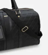 Bolso tipo valija grande color negro, con textura geométrica en relieve, cierre de cremallera, correa de mano, asa larga desmontable y tag de identificación.