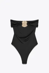 Bañador negro tipo bandeau de escote recto y drapeado, con aplicación de pieza metálica circular dorada en el pecho y forro interior.