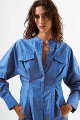 Camisa de manga larga color azul con finas rayas verticales blancas. Presenta cuello mao, botones frontales y dos bolsillos de parche con tapa en el pecho. La prenda tiene un diseño ajustado en la cintura con pinzas y un volumen holgado en las mangas.