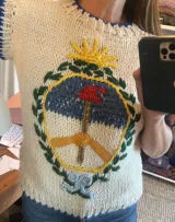 Chaleco tejido de lana merino color crudo, con cuello redondo y sisas ribeteadas en azul. Presenta bordado del escudo de Argentina en el frente.