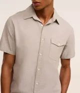 Camisa de hombre de manga corta en lino color blanco, con cuello clásico, botonadura frontal y bolsillo de parche en el pecho. Corte recto.