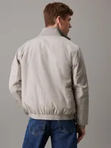 Campera Calvin Klein color gris claro, confeccionada en nailon 100% reciclado. Tiene cuello alto de canalé, puños y bajo elásticos, cierre de cremallera, bolsillos en la cintura, forro ligeramente acolchado y logo de la marca en el pecho y la manga.