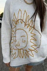 Buzo de tejido de punto color crudo, con cuello redondo y manga larga. Presenta un bordado frontal grande con un diseño abstracto que simula un rostro con rayos solares.