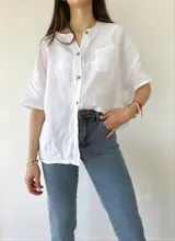 Camisa de lino y algodón color crudo, de corte recto, manga corta y cuello redondo con abertura frontal con botones. Tiene un bolsillo en el pecho con detalle de puntilla.