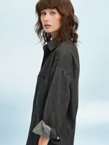 Camisa de denim negro con corte oversize, cuello clásico, cierre frontal con botones metálicos y bolsillo de parche en el pecho.