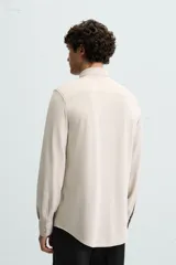 Camisa color beige de corte slim fit, confeccionada en tejido elástico con cuello italiano abierto, manga larga con puños abotonados y cierre frontal con botones.