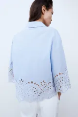 Camisa celeste de popelina con cuello solapa y manga ancha por debajo del codo. Bajo acabado con bordados perforados. Cierre frontal con botones.