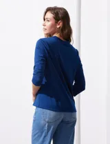 Blusa blanca de poliéster y elastano, con cuello a la base, escote en V y mangas tres cuartos. Presenta dos bolsillos frontales con cierre metálico dorado.