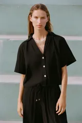 Camisa negra de manga corta con cuello solapa y escote pico. Detalle de nido de abeja en la espalda y cierre frontal con botones.