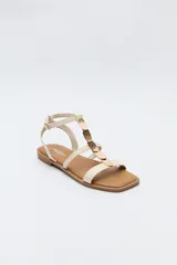 Sandalias planas color beige con tiras finas y aplique metálico central. Cierre con hebilla lateral y base marrón.