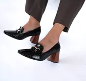 Zapato mocasín de cuero negro con estampado brillante y detalle metálico dorado en la parte delantera. Tiene taco geométrico escultural color madera.